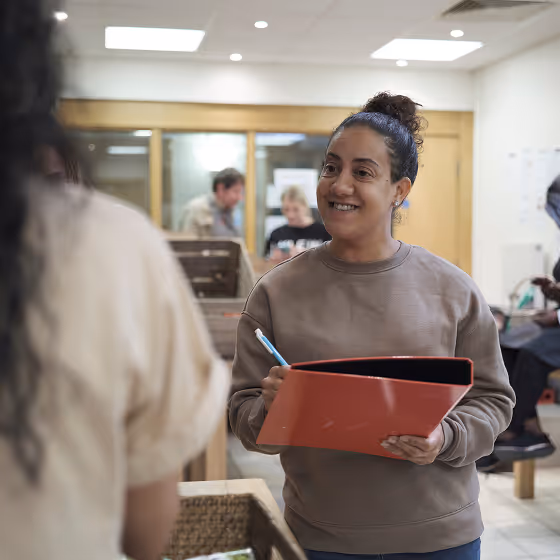 Woman holding a red folder and pen, smiling and talking to another person in a casual indoor setting.
