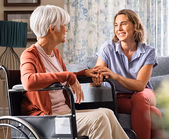Young woman holding hands and smiling with an elderly woman in a wheelchair in a living room.