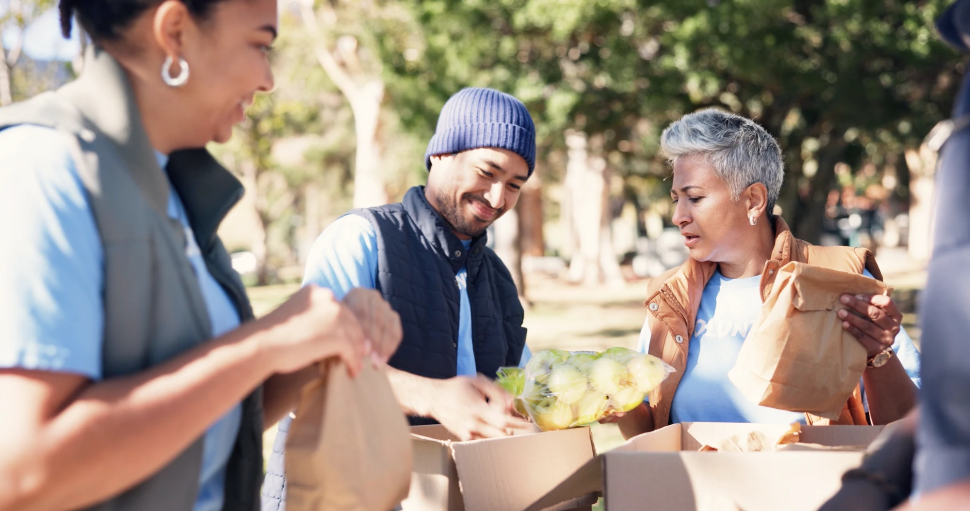 Three volunteers packing food items into bags outdoors in a park.