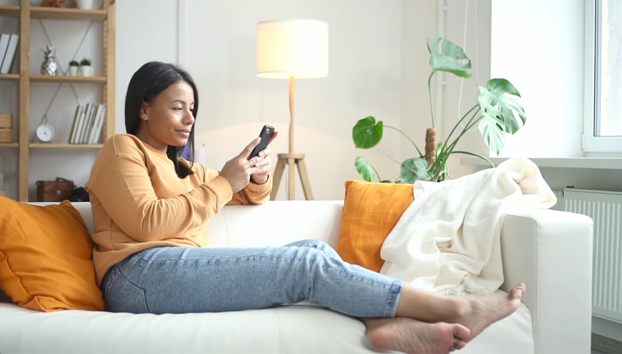 Woman in a mustard sweater and jeans sitting barefoot on a white couch, looking at her smartphone with orange pillows and a white blanket nearby.