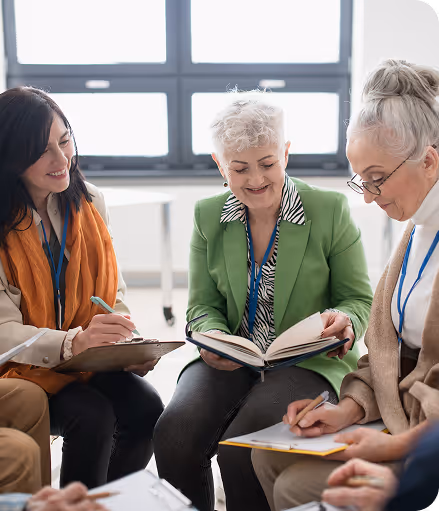 Three older women sitting indoors, one reading from a book while the others write on clipboards.