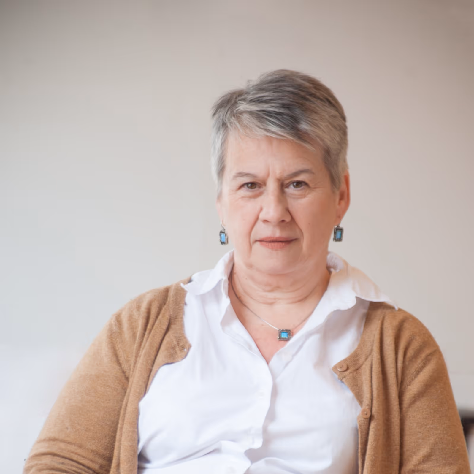 Middle-aged woman with short gray hair wearing a white shirt and brown cardigan with turquoise jewelry, sitting against a plain light background.