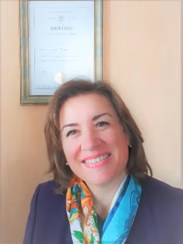Smiling woman with shoulder-length brown hair wearing a colorful scarf and dark blazer, with a framed diploma on the beige wall behind her.