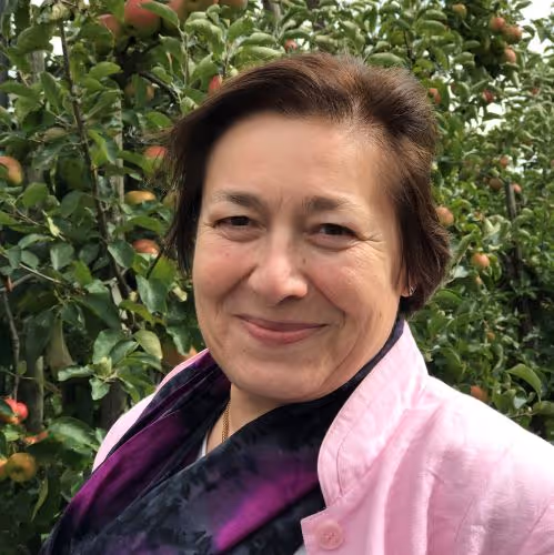 Smiling woman in a light pink jacket and dark scarf standing outdoors in front of leafy green trees with apples.