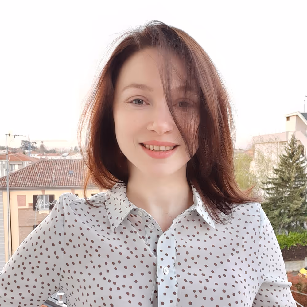 Smiling young woman with shoulder-length brown hair wearing a white shirt with brown polka dots, posing outdoors with buildings and trees in the background.