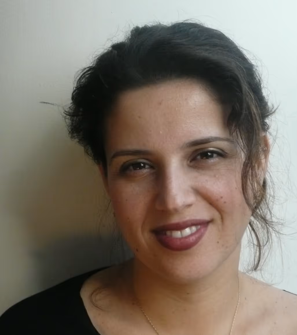 Smiling woman with dark hair pulled back, wearing a black top and light makeup, against a plain light background.
