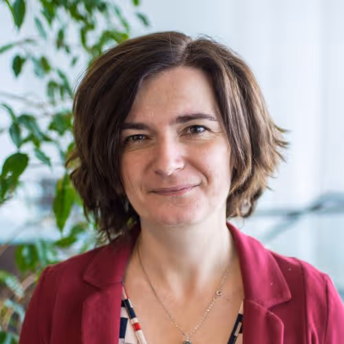 Portrait of a woman with short brown hair wearing a red blazer and a patterned top, smiling softly against a blurred indoor background with green plants.