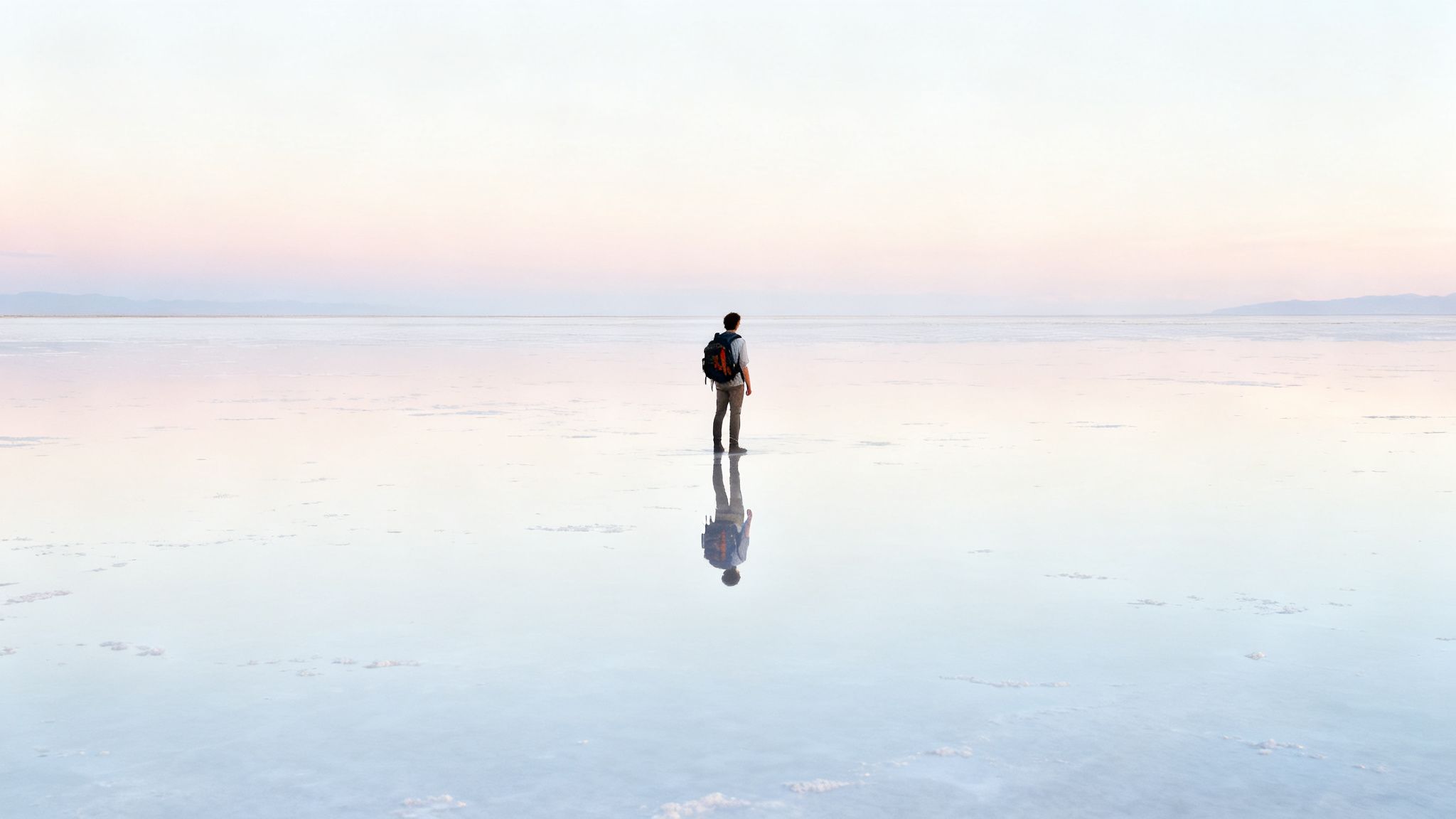 A lone traveler with a backpack stands on a vast, reflective salt flat under a pastel sky.