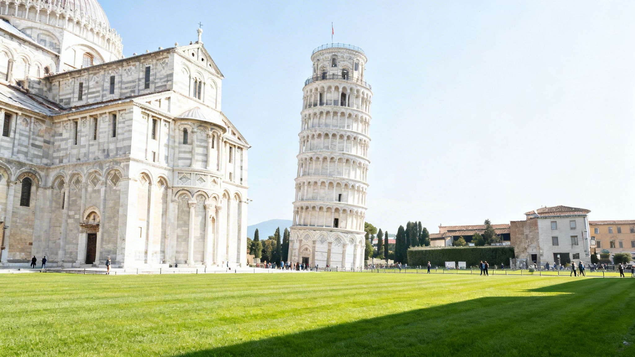 The iconic Leaning Tower of Pisa and Pisa Cathedral stand tall on a sunny day with green grass.