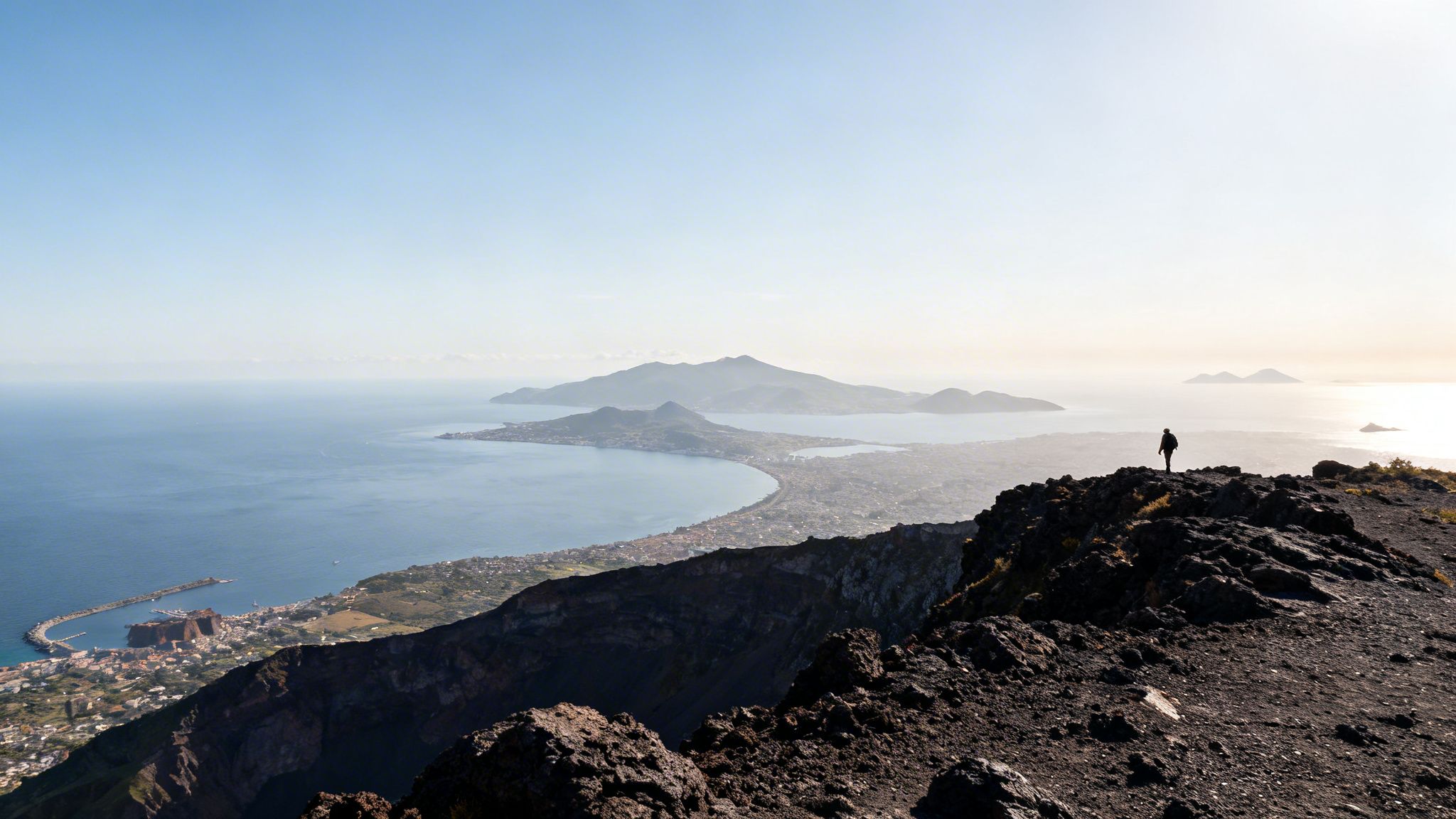 A lone figure on a volcanic peak overlooks a scenic bay, coastal town, and distant islands.