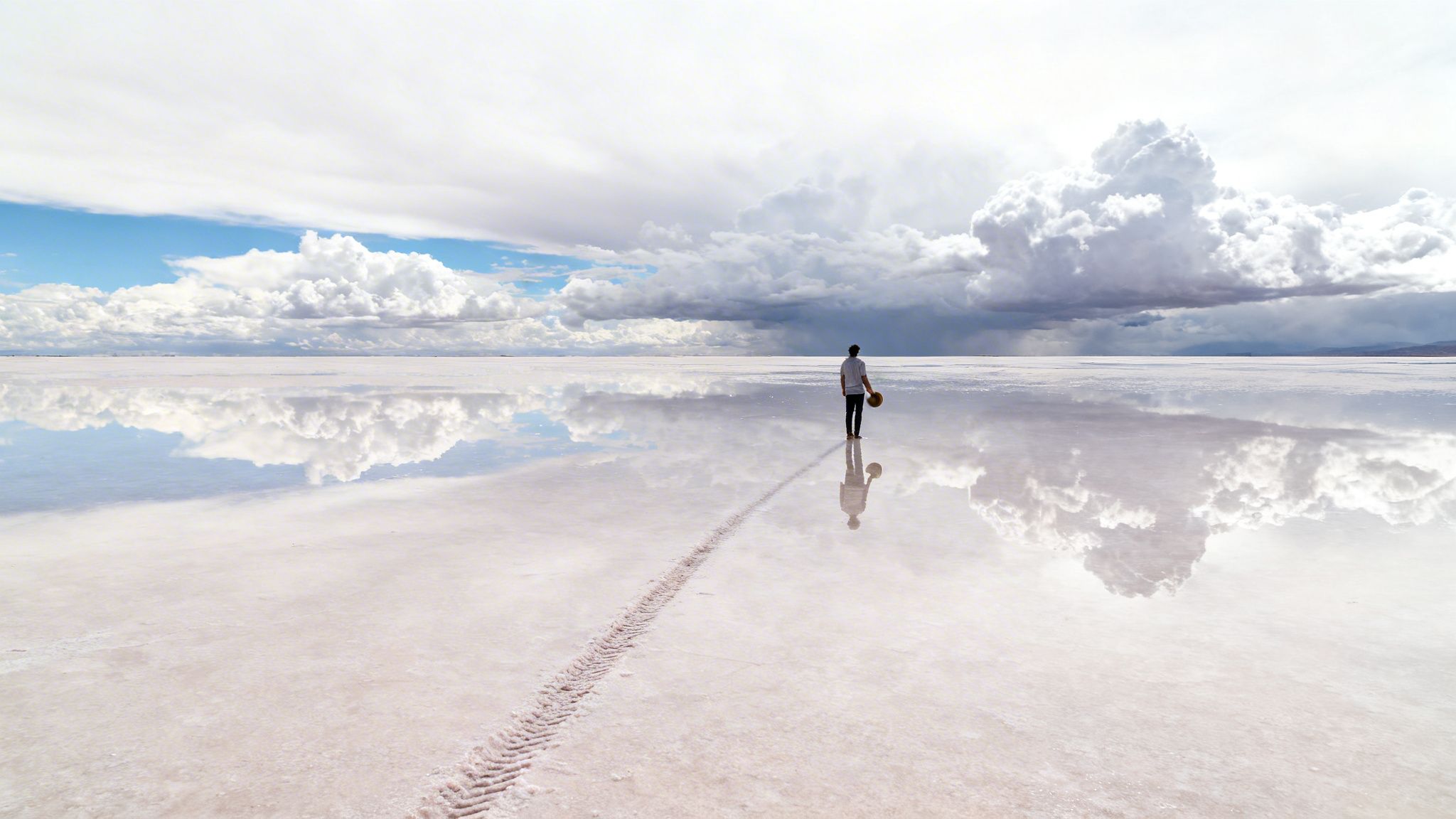 A person stands on a vast, reflective salt flat under a cloudy sky, with tire tracks leading away.