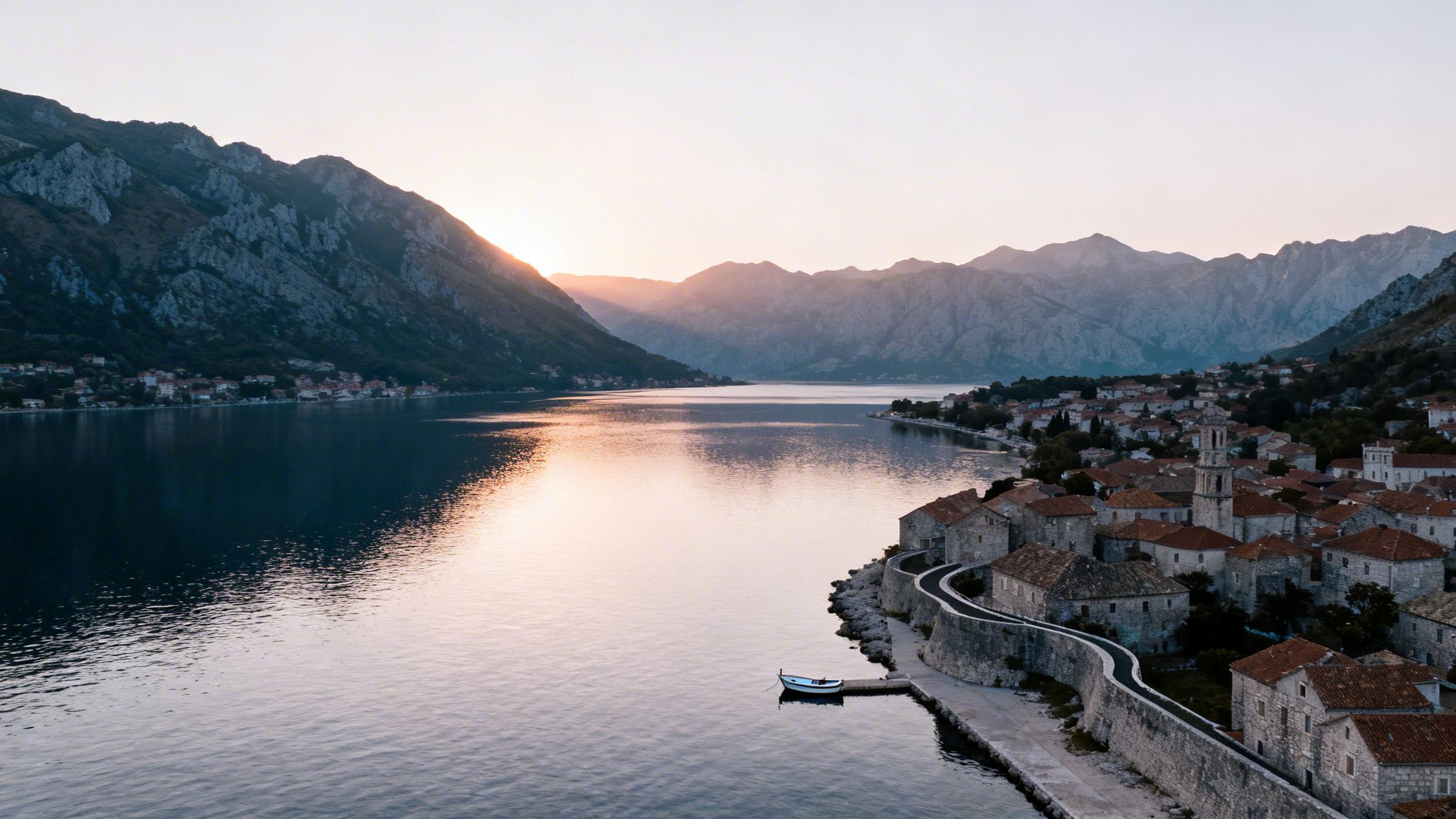 Panoramic view of a stunning coastal town with red-roofed buildings nestled by a serene bay at sunset.