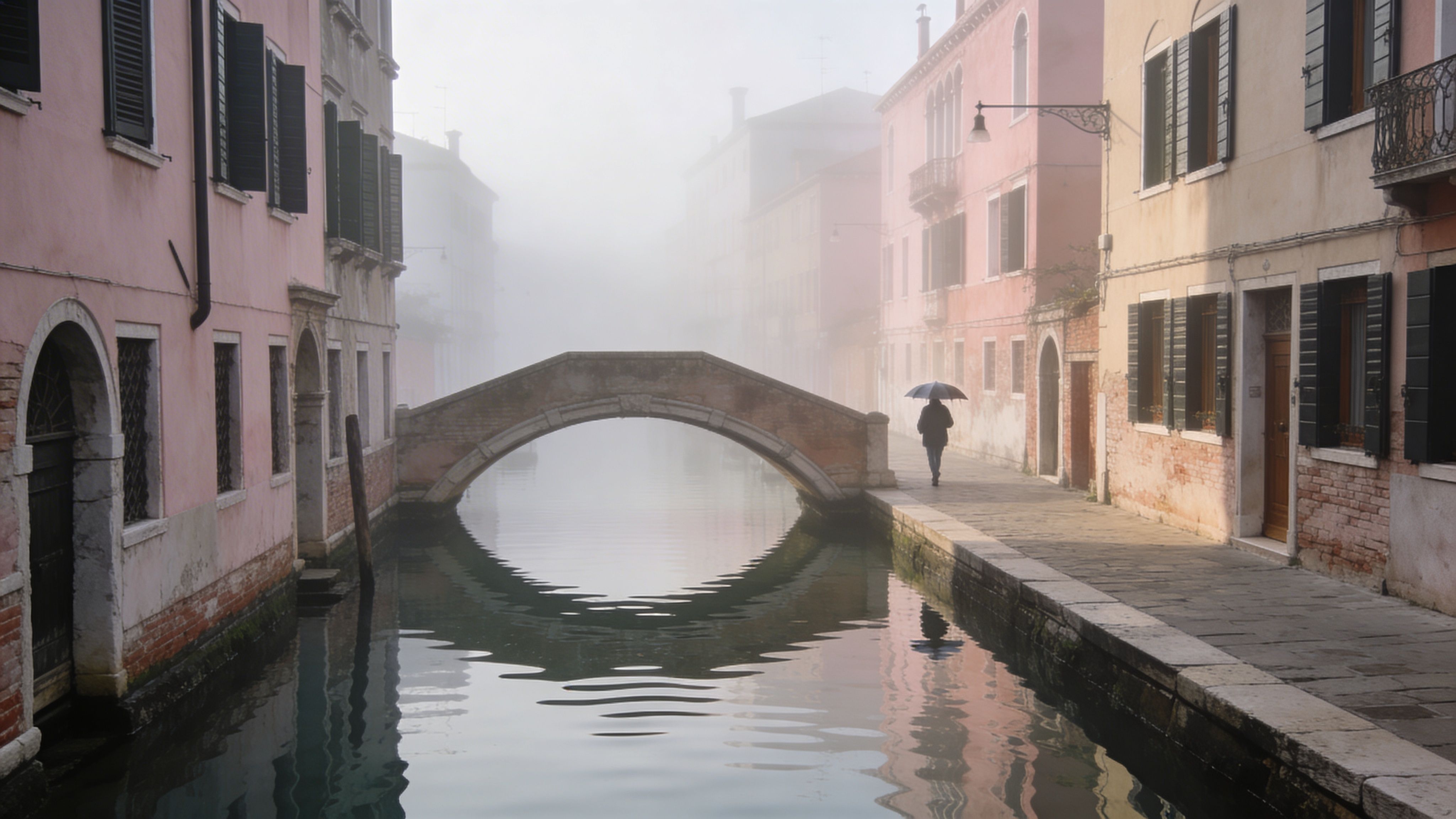 A person walking with an umbrella across a quiet stone bridge in foggy Venice during the day.