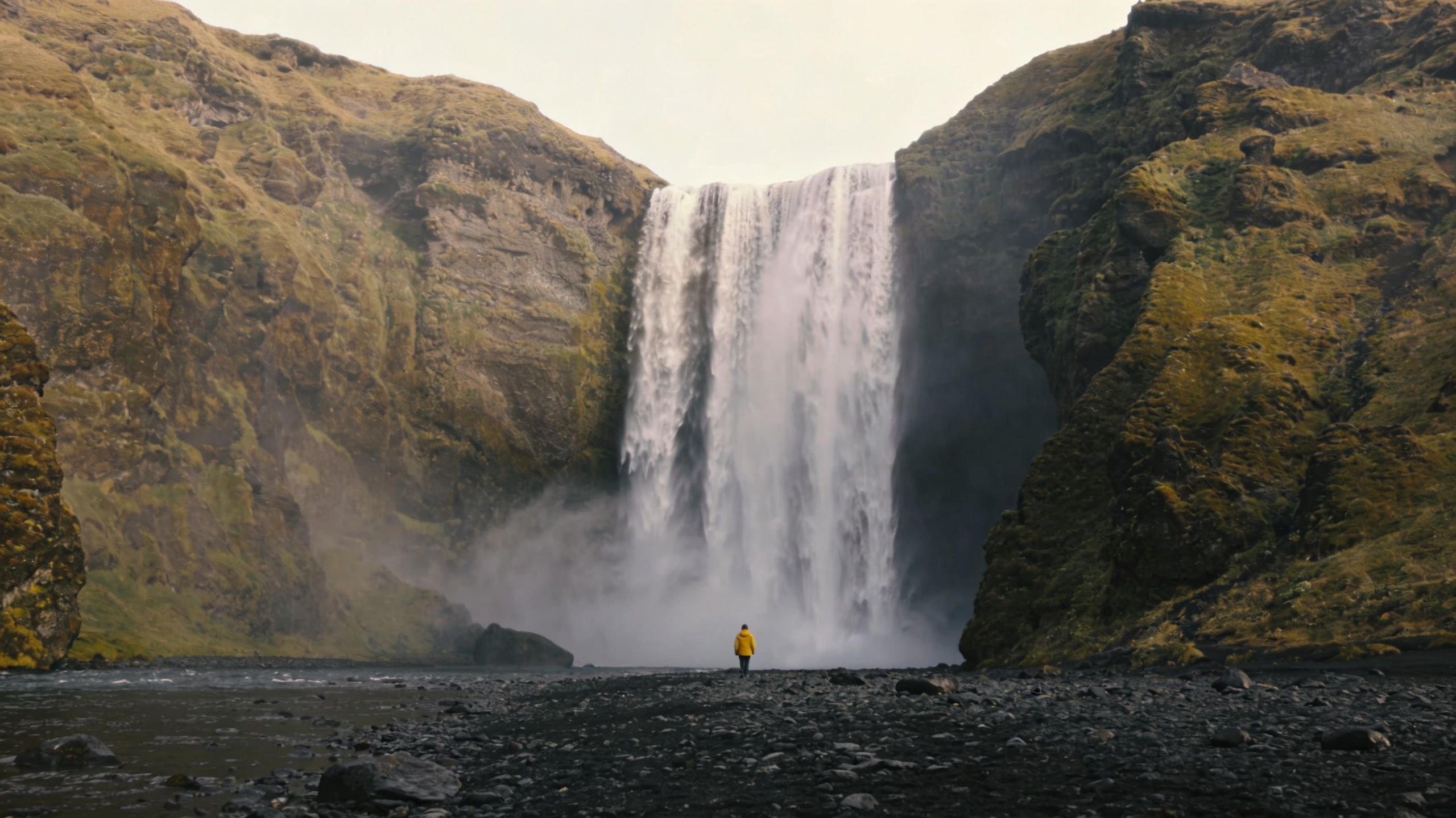 A person in a yellow jacket stands before the majestic Skogafoss waterfall in Iceland