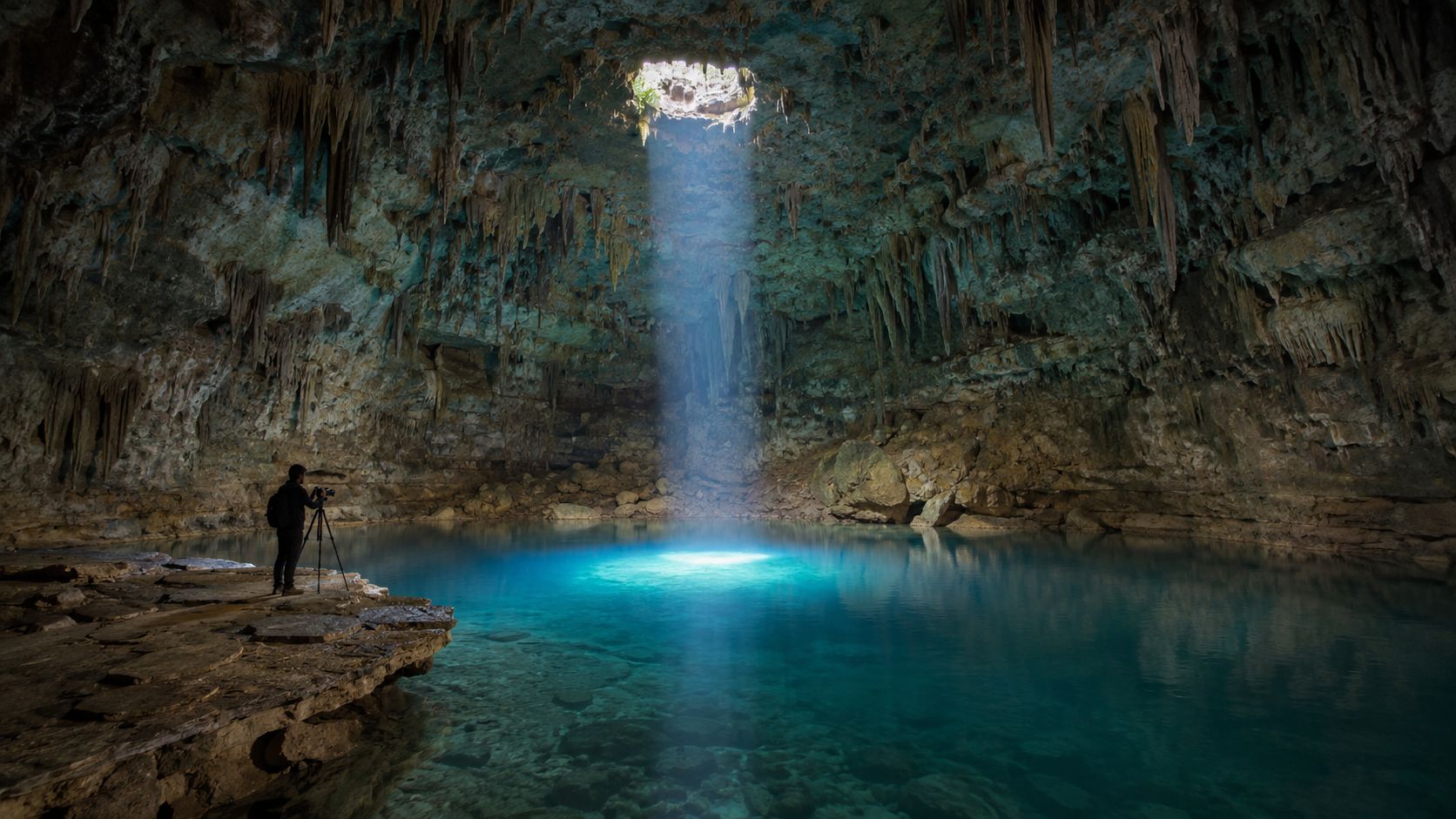 A photographer with a tripod stands inside a beautiful cenote with a light beam illuminating blue water.