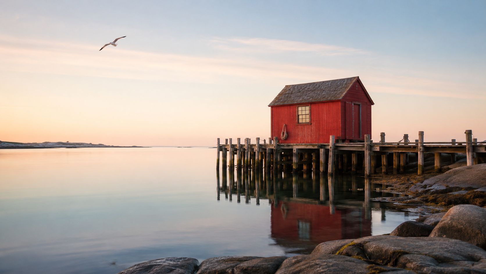 A serene red wooden cabin sits on a wooden pier over calm water at sunset in Maine.