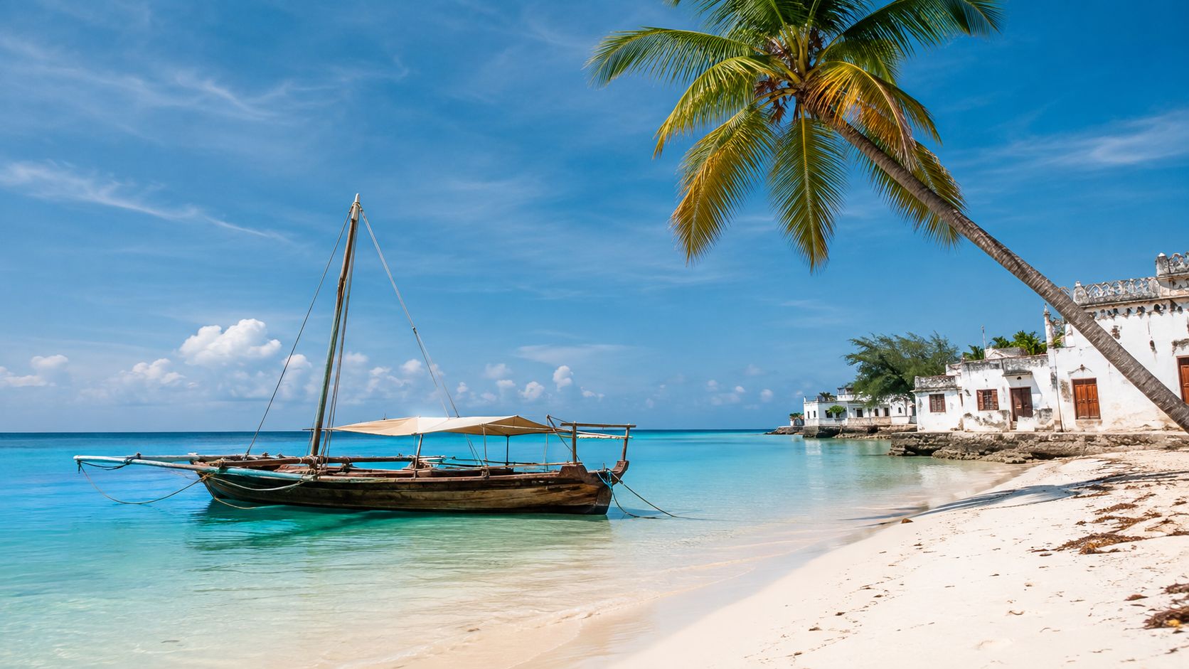 A traditional wooden dhow boat floating on turquoise tropical water near a sandy beach and white buildings.
