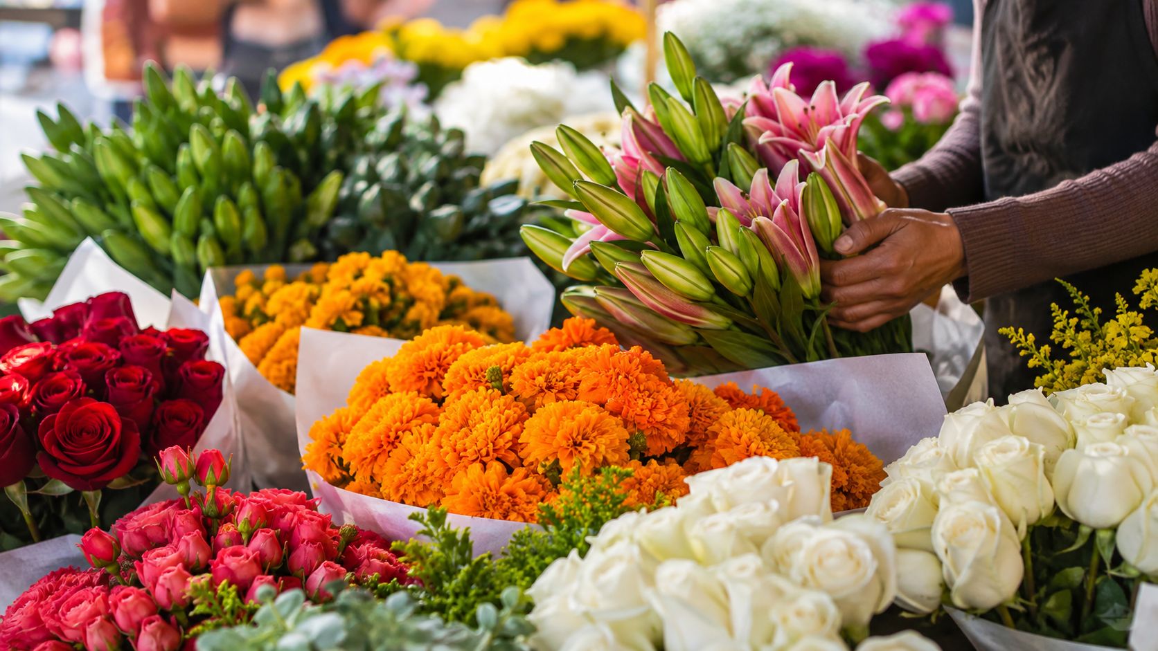 A person arranging a fresh bouquet of pink lilies at a vibrant flower market in Mexico City.