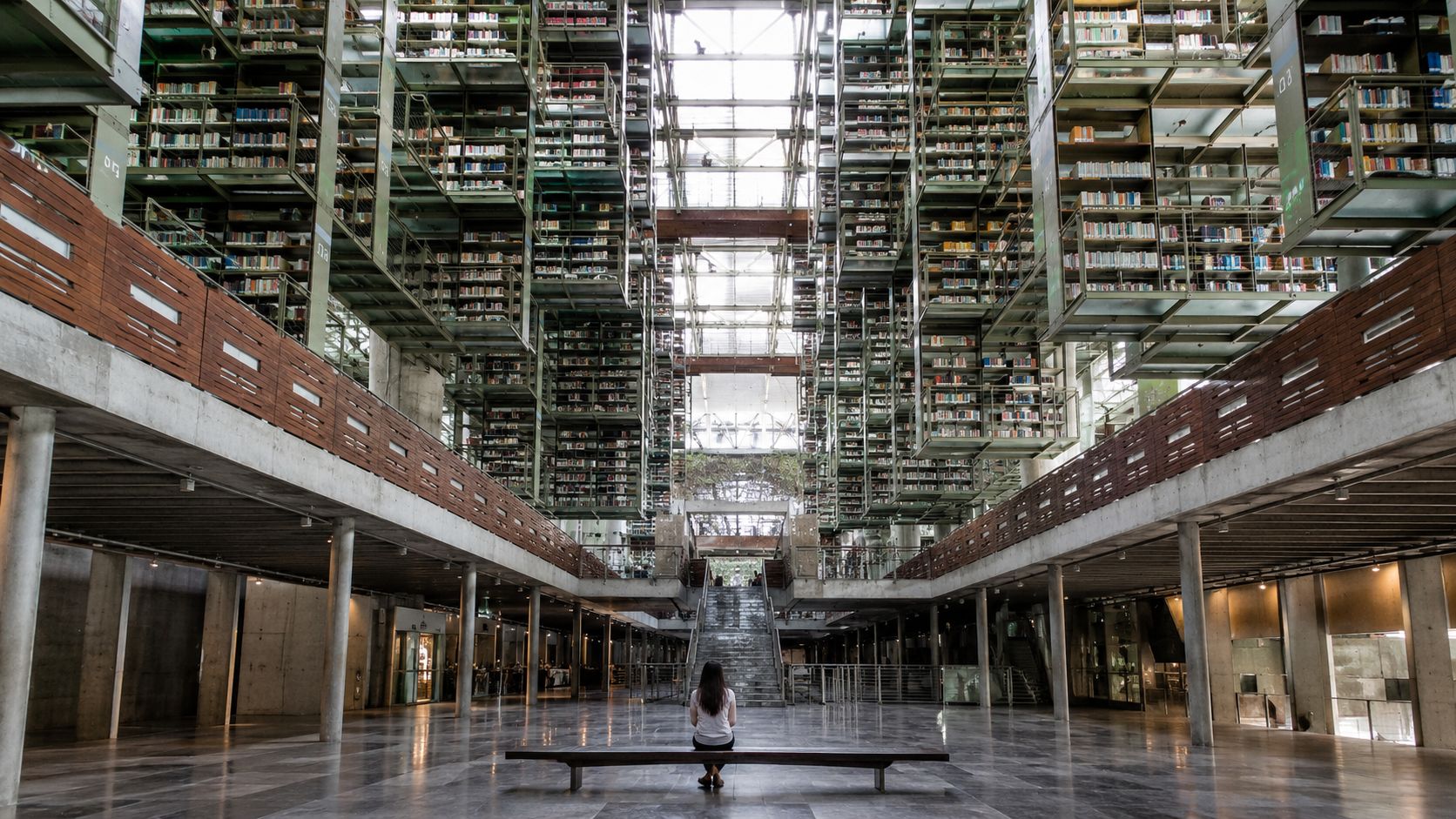 A woman sitting alone on a wooden bench inside the stunning, multi-level Vasconcelos Library in Mexico City.