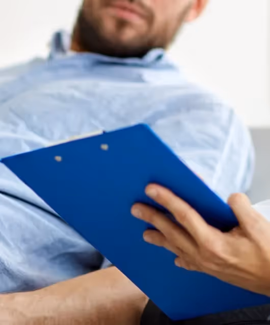 Male patient holding a blue medical clipboard while sitting on an examination table in a clinic.