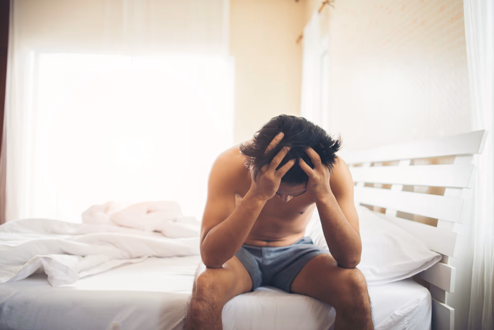 Man sitting on the edge of a bed in a bright room with his head in his hands.