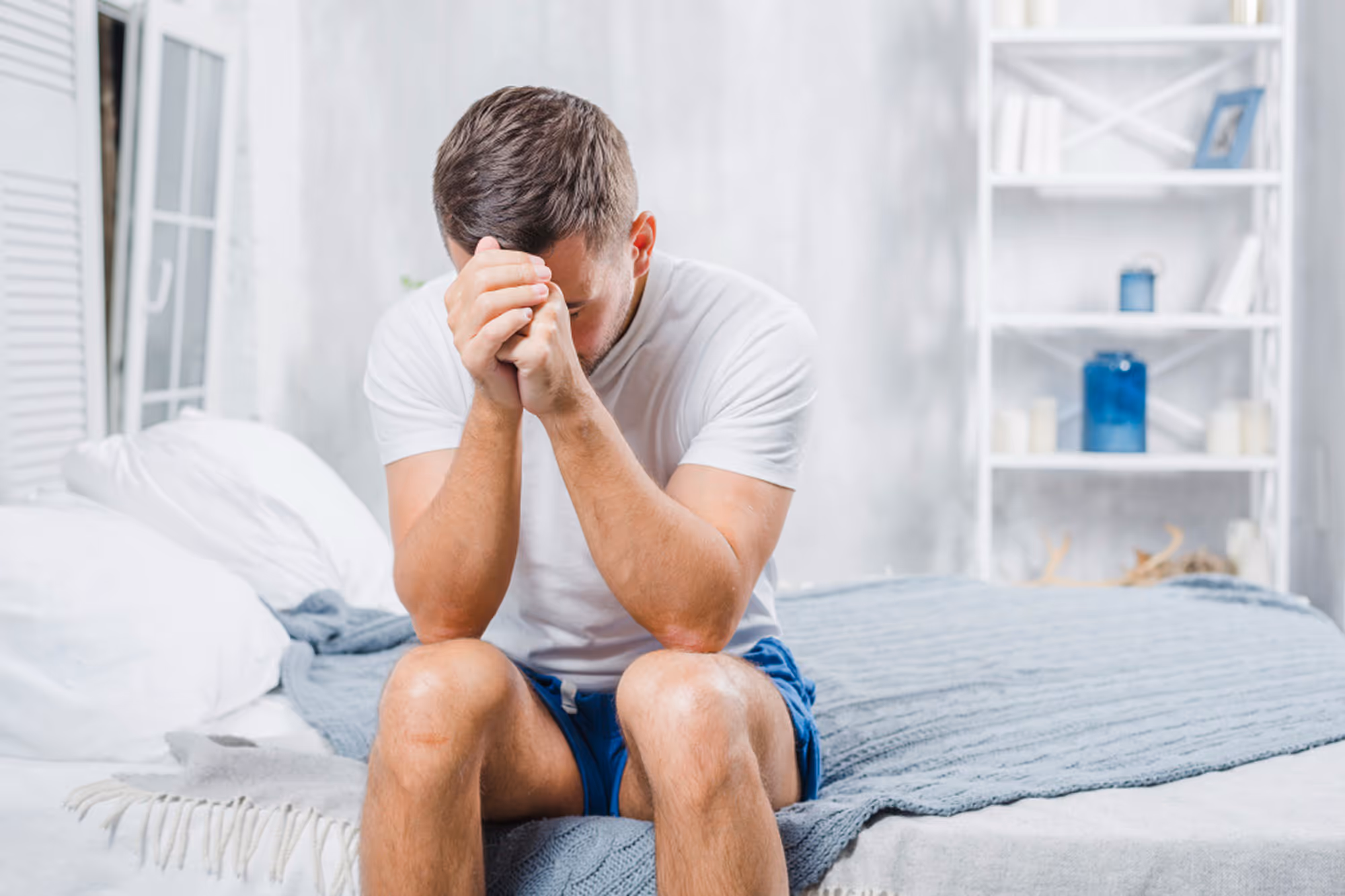 Man sitting on the edge of a bed with his head in his hands in a bedroom.