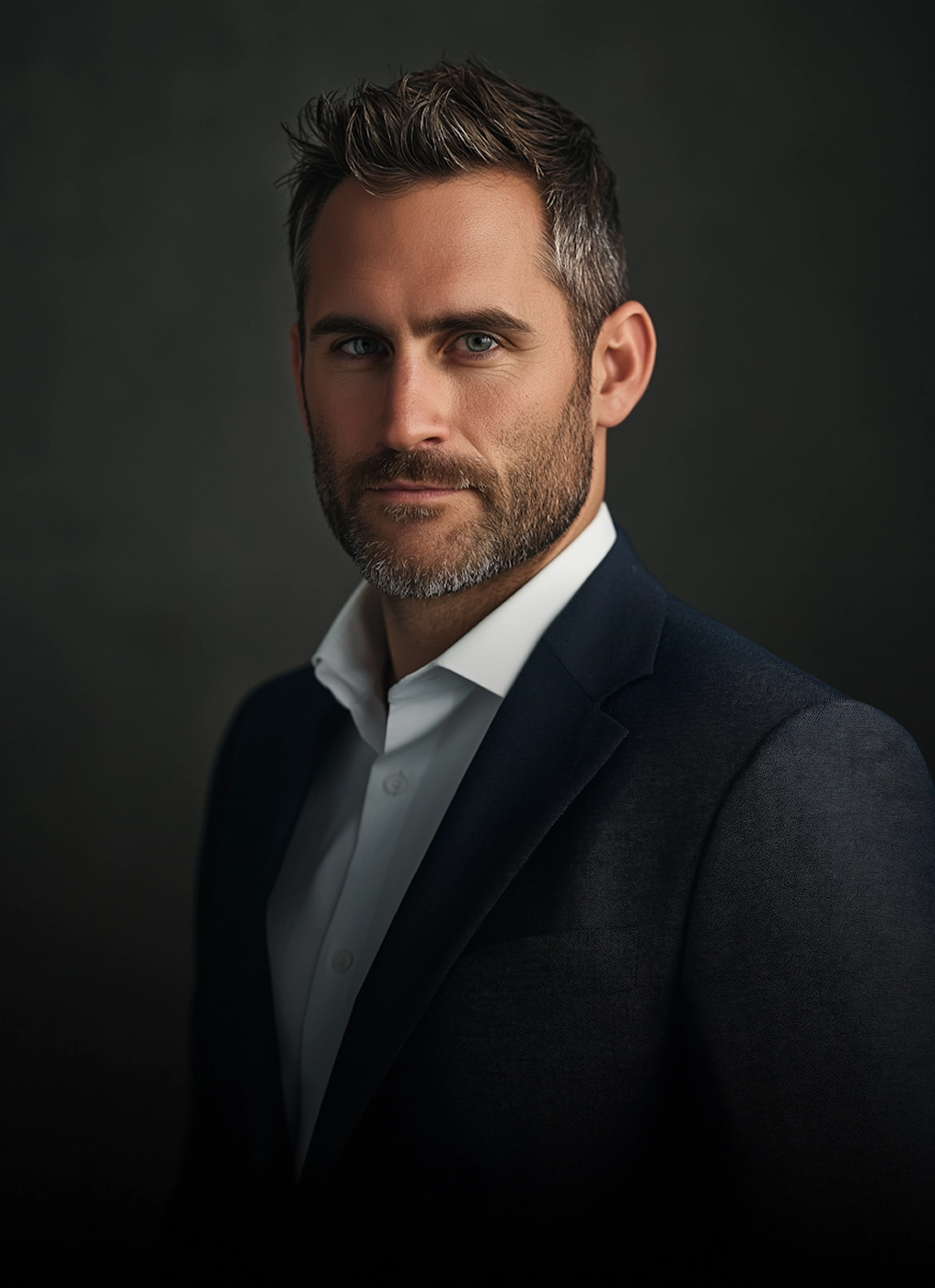 Portrait of a confident man with short dark hair and beard wearing a white shirt and dark blazer against a dark background.