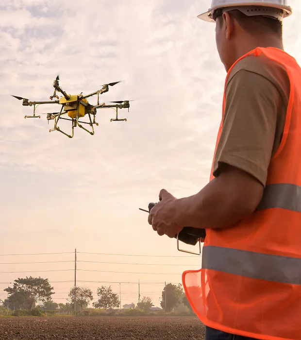 Un télépilote en gilet orange et casque contrôlant un drone agricole jaune au-dessus d’un champ au crépuscule.