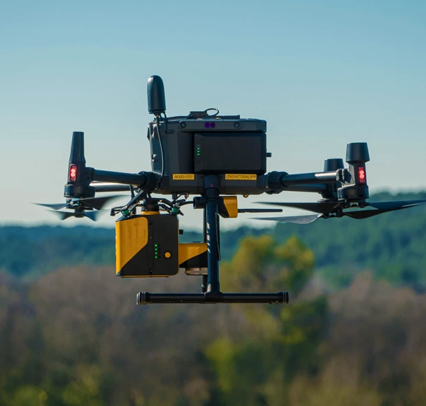 Drone noir équipé de capteurs jaunes volant en extérieur avec un ciel bleu et des arbres flous en arrière-plan.