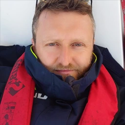 Portrait d'un homme avec une barbe claire portant une veste marine et une écharpe rouge.