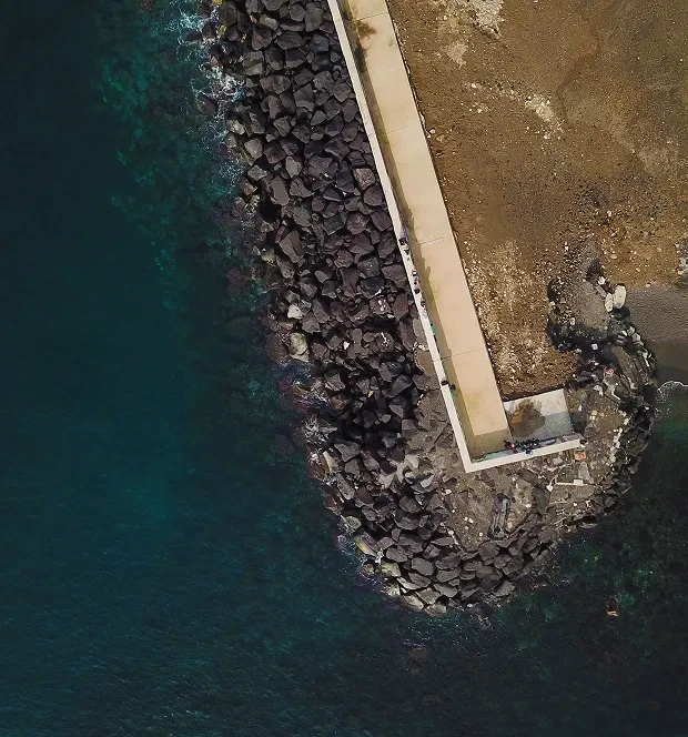 Vue aérienne d'une digue en béton bordée de rochers sombres entre une mer bleu foncé et une terre sèche.