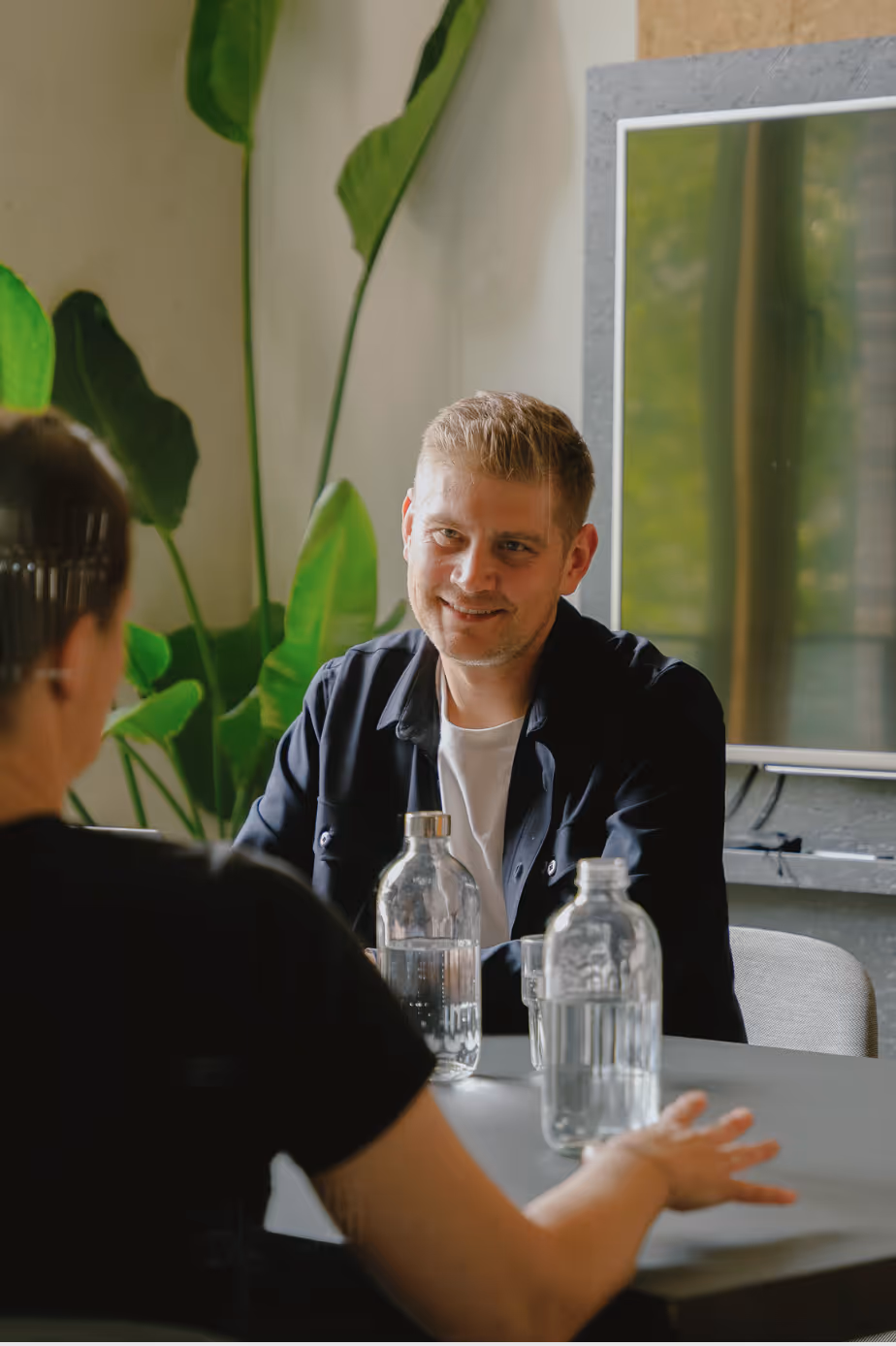 Zwei Personen sitzen sich an einem Tisch gegenüber, einer lächelt, im Hintergrund große grüne Pflanzen.