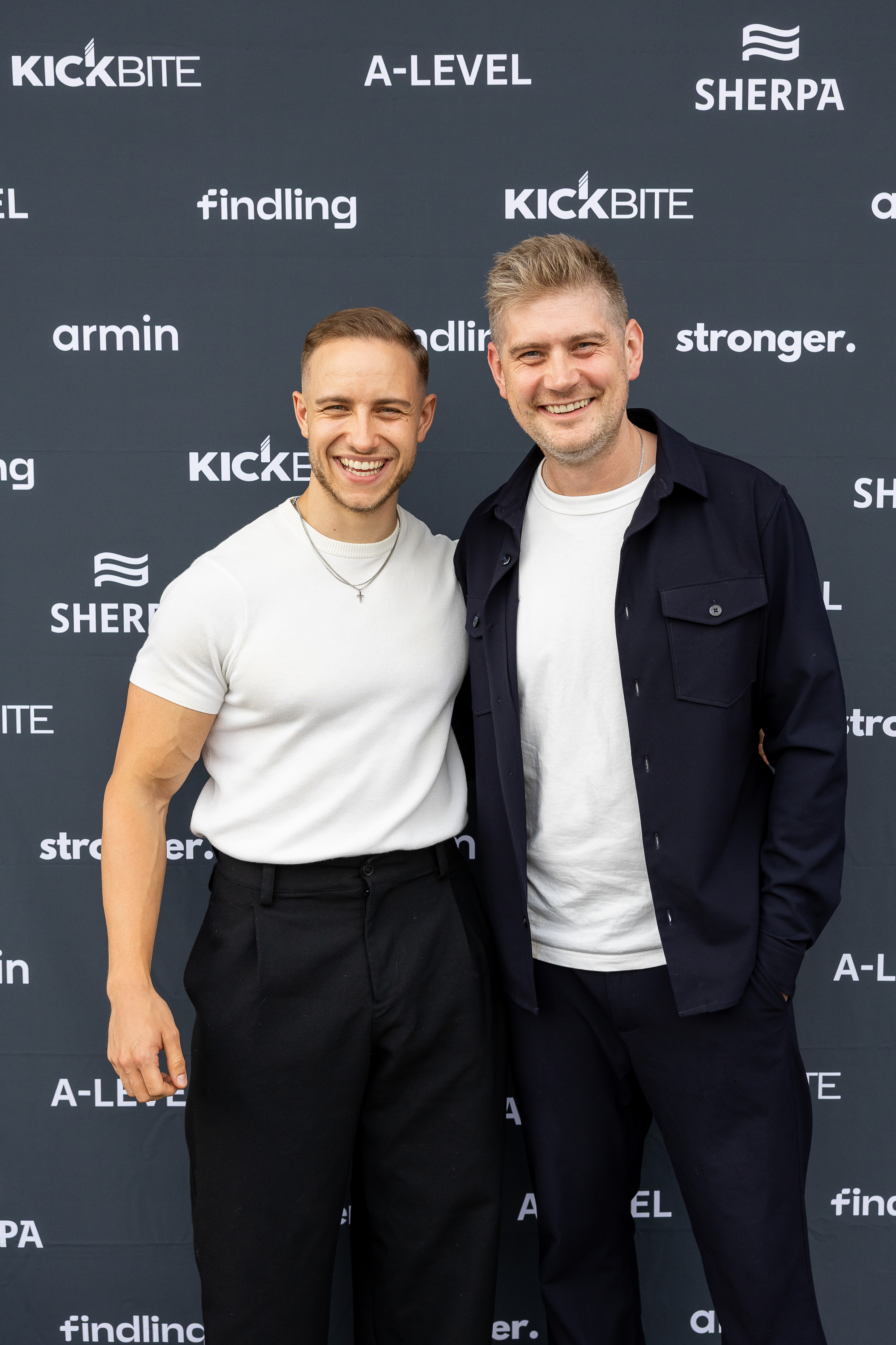 Zwei lächelnde Männer stehen vor einem dunklen Hintergrund, einer trägt ein weißes T-Shirt und der andere ein weißes T-Shirt mit einer dunklen Jacke.