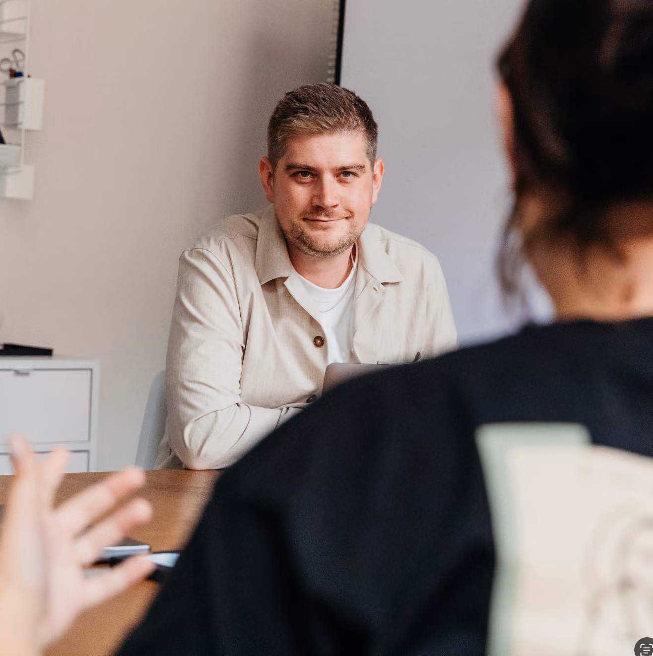 Mann mit beige Hemd sitzt an einem Tisch und hört einer anderen Person zu, die im Vordergrund unscharf zu sehen ist.