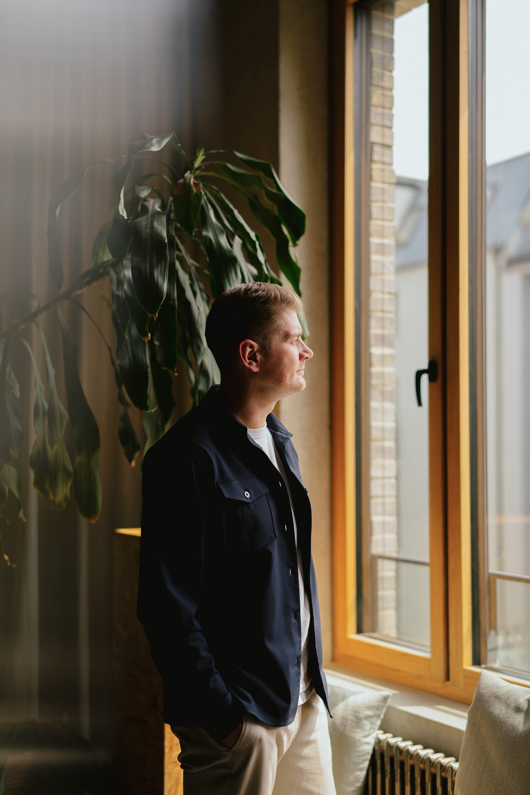 Ein Mann mit dunkler Jacke blickt nachdenklich aus einem großen Fenster, im Hintergrund eine grüne Zimmerpflanze.