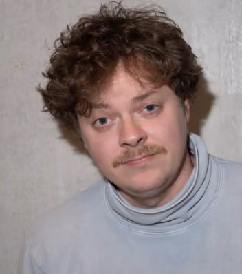 Mann mit krausem braunem Haar, Schnurrbart und grauem Rollkragenpullover vor einer grauen Wand.
