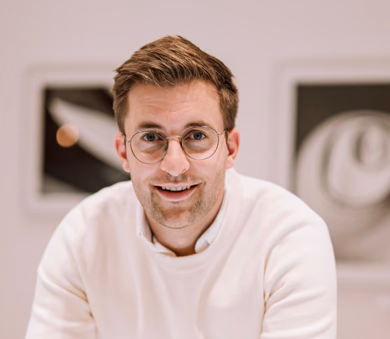 Junger Mann mit Brille und weißem Pullover lächelt freundlich in die Kamera.