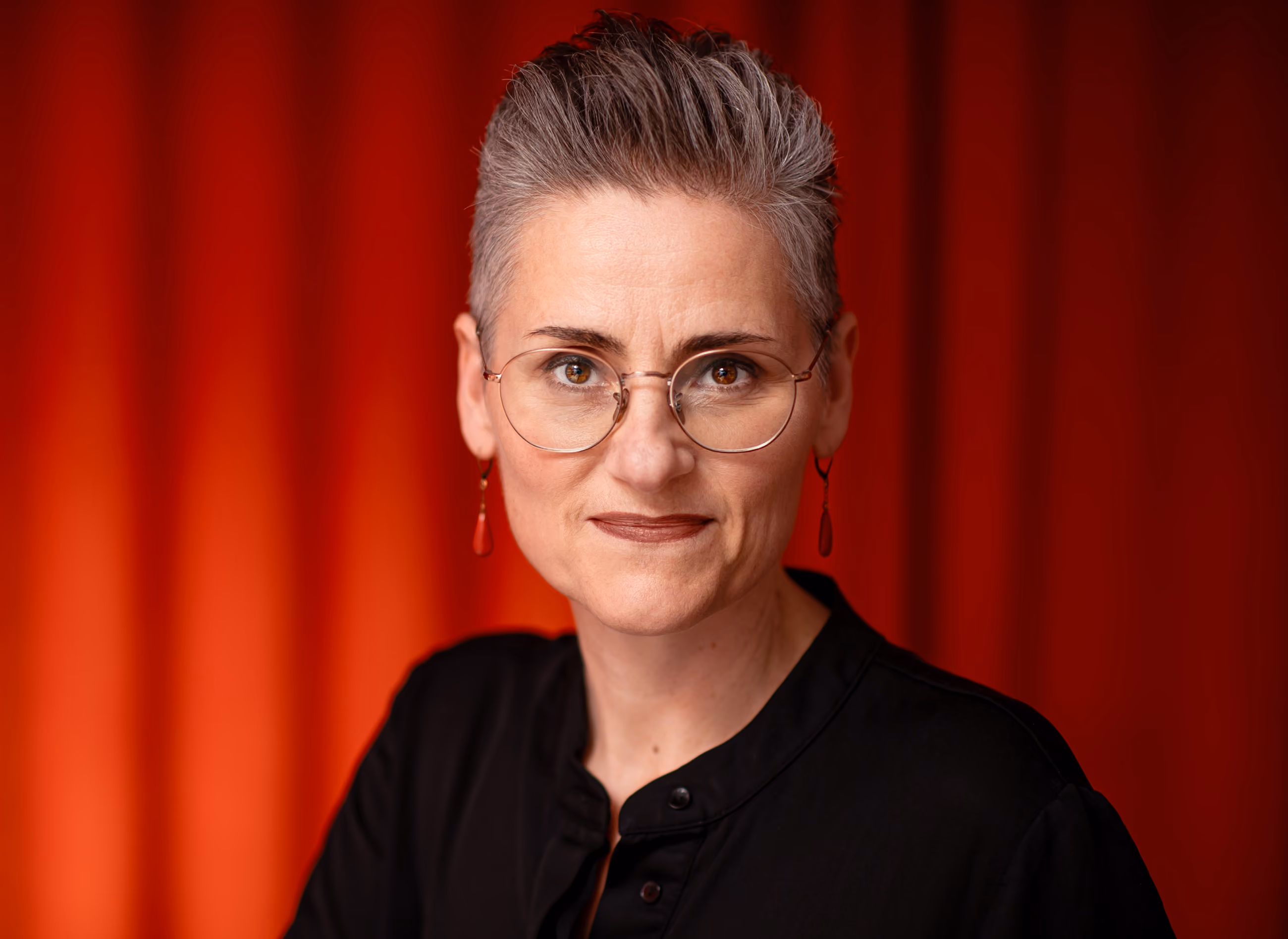 Porträt einer Frau mit kurzen grauen Haaren, runden Brillengläsern und roten Ohrringen vor rotem Hintergrund.