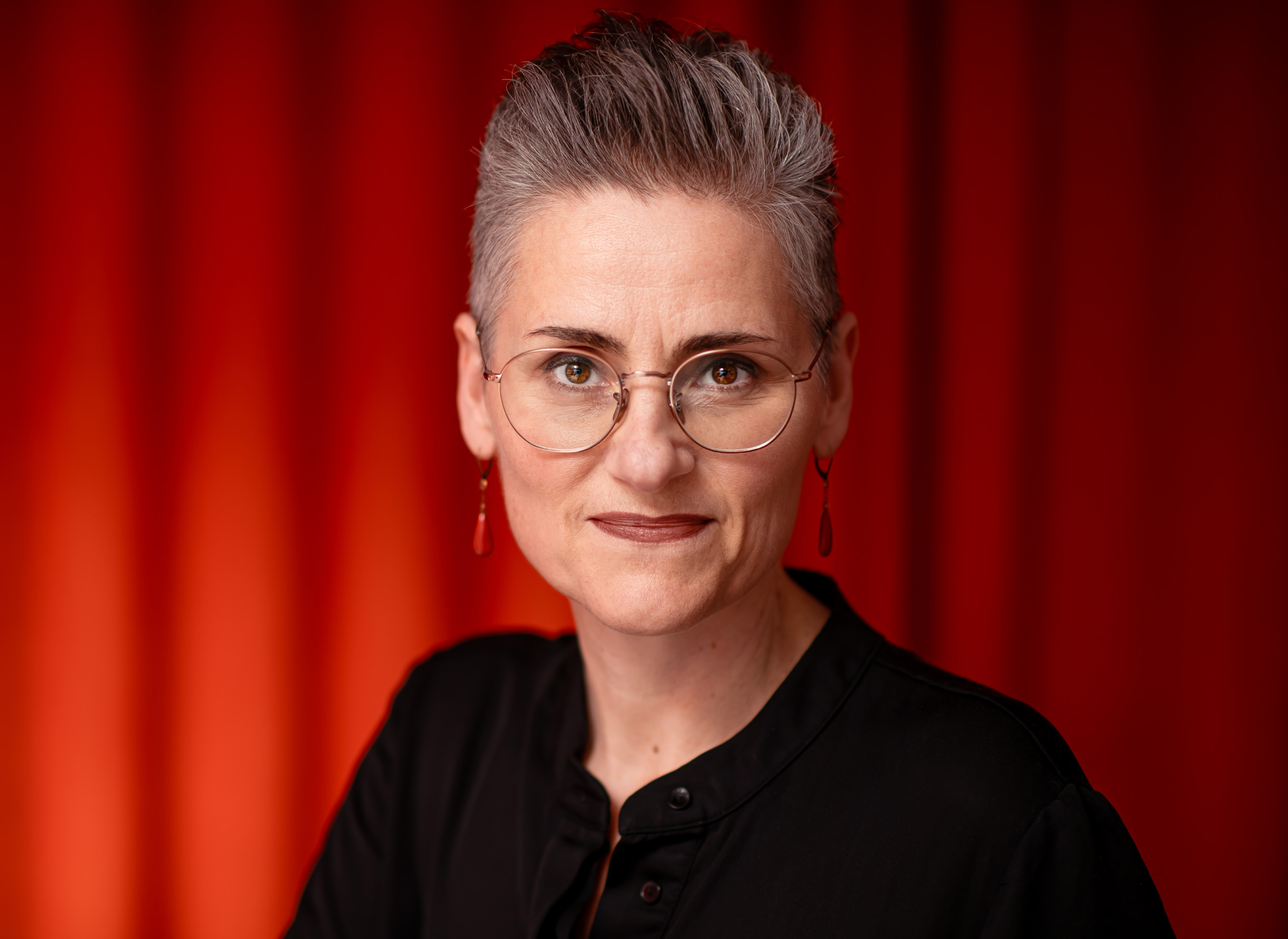Porträt einer Frau mit kurzen grauen Haaren, runden Brillengläsern und roten Ohrringen vor rotem Hintergrund.