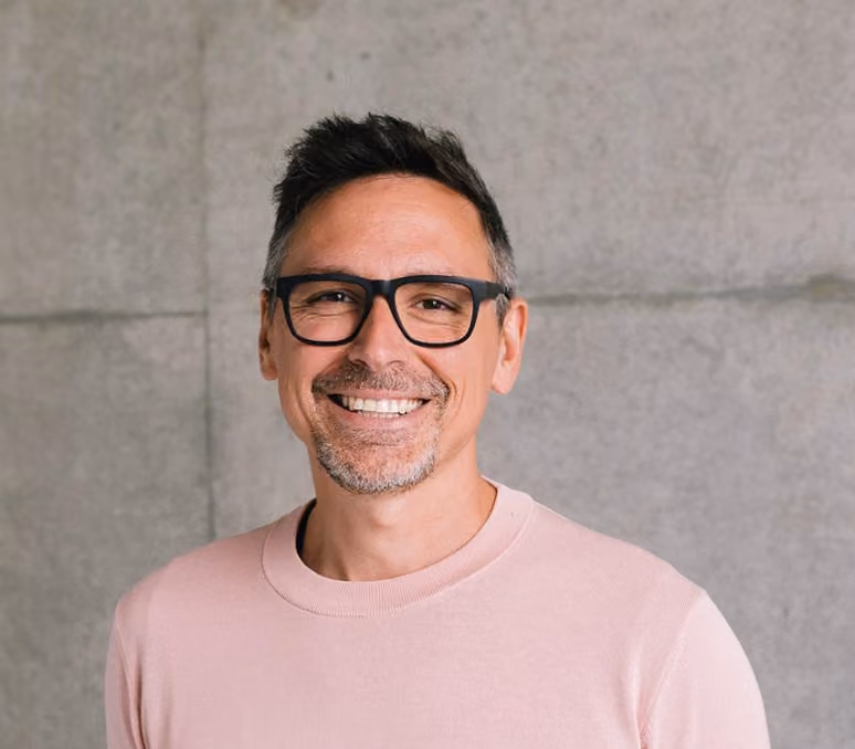 Lächelnder Mann mit dunklen Haaren, Brille und rosa Pullover vor einer grauen Wand.
