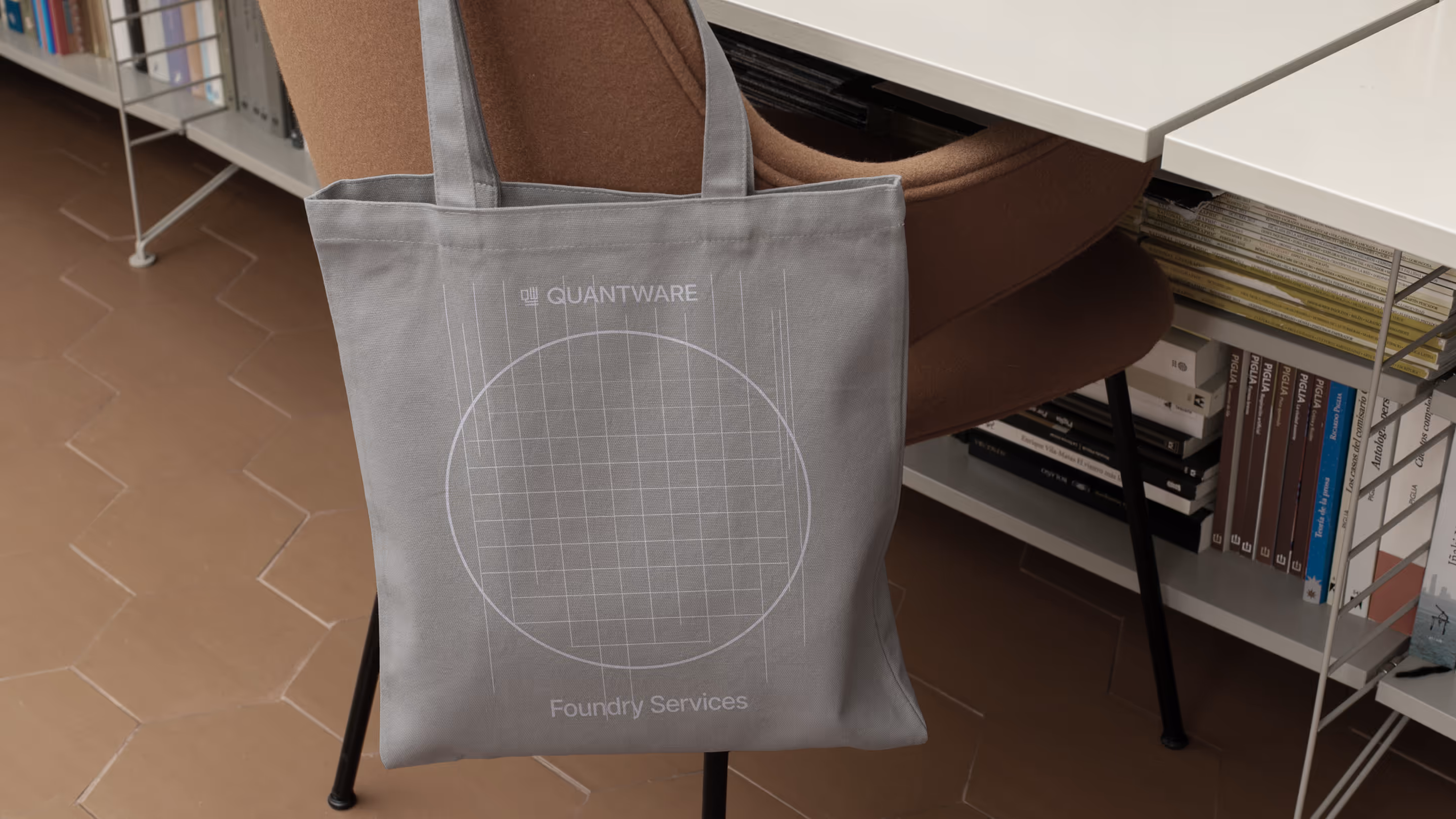 Gray tote bag with a circular grid design and the text 'QUANTWARE Foundry Services' hanging on a chair in a room with tiled flooring and bookshelves.