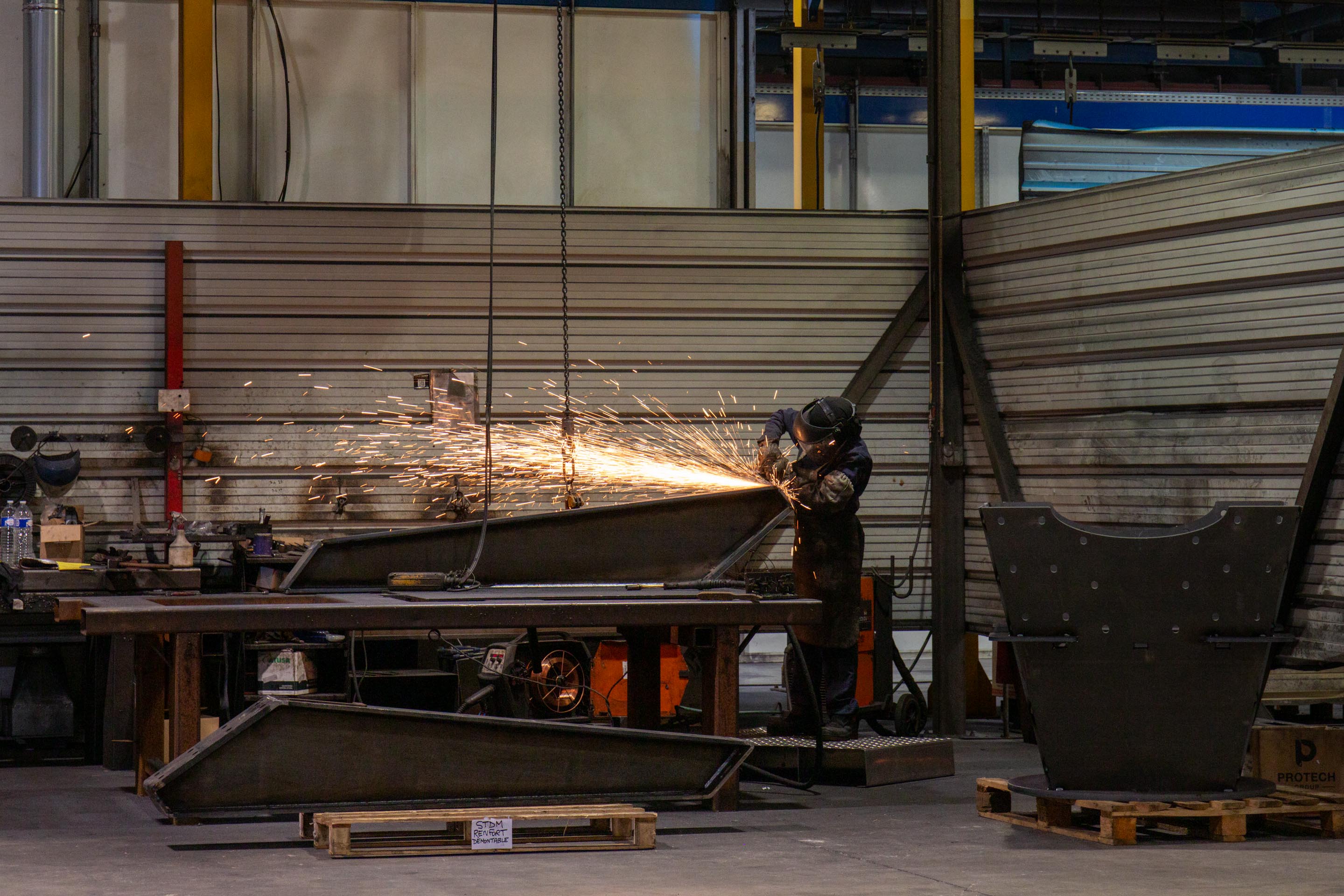 Soudeur portant un casque de protection qui travaille sur une pièce métallique avec une meuleuse dans un atelier industriel.