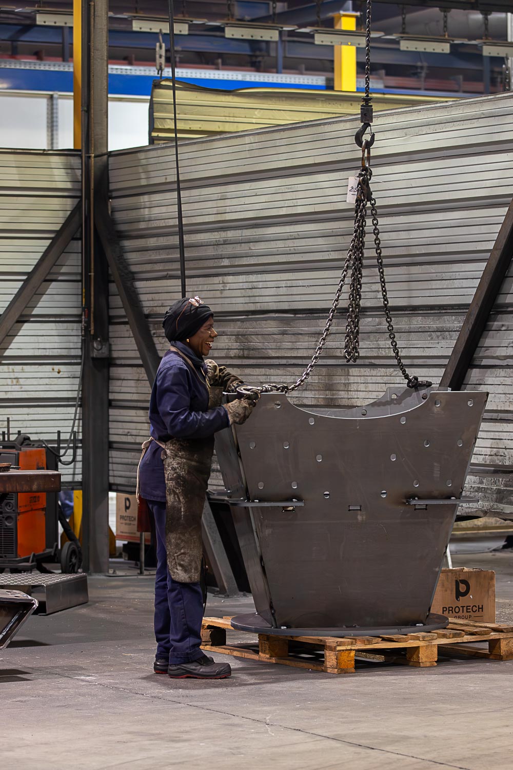Femme en bleu de travail et munie de ses équipements de protection individuelle, manipulant une grosse pièce métallique suspendue, dans un atelier industriel.