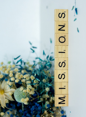 Wooden Scrabble tiles spelling the word "MISSIONS" stacked vertically next to a bouquet of dried flowers with blue, white, and yellow hues.