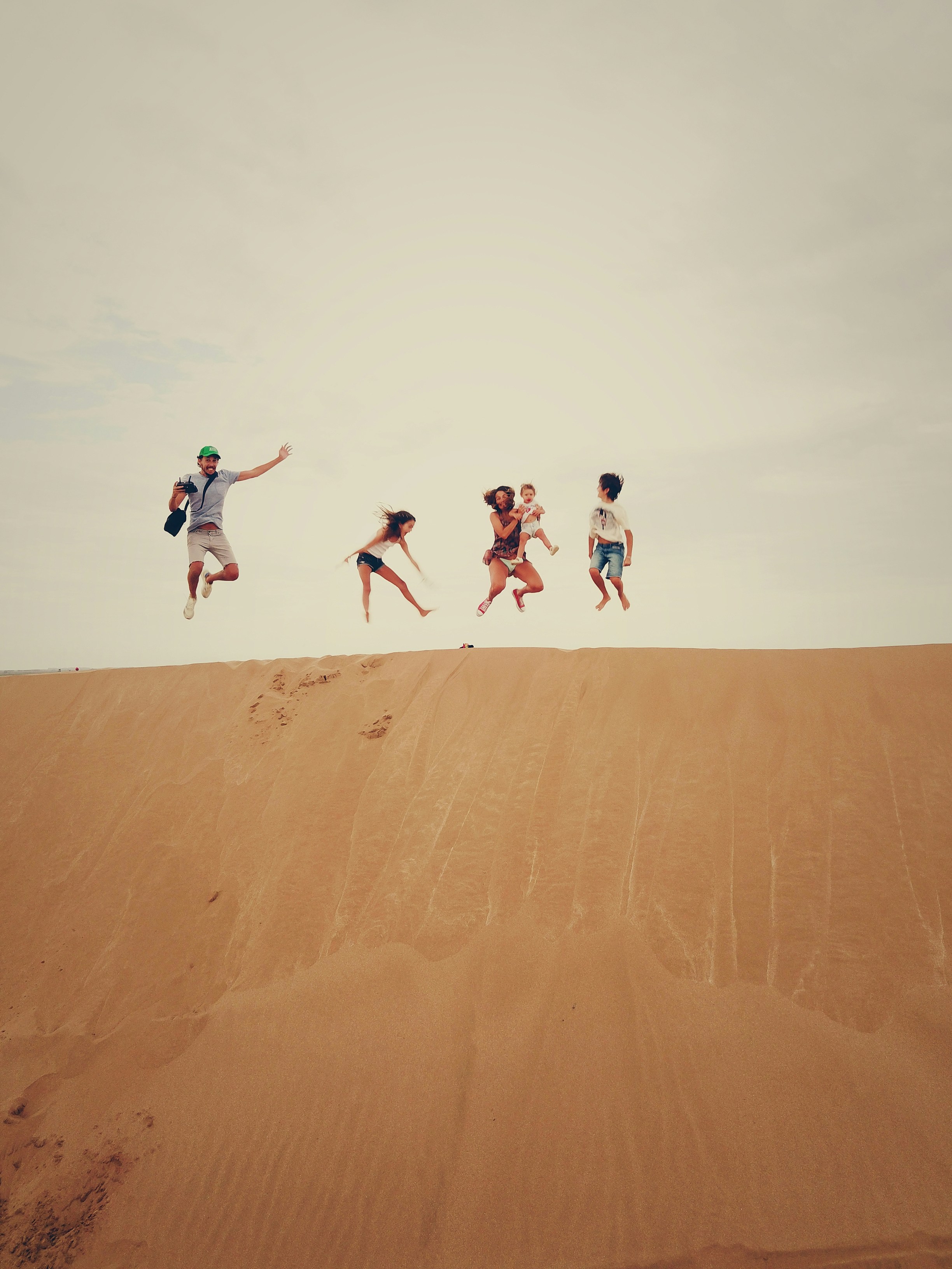 Five people jumping in the air on a sand dune against a cloudy sky.