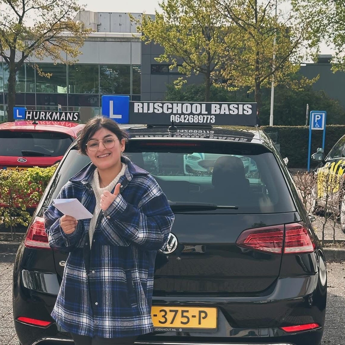 Jonge vrouw staat voor een zwarte auto van rijschool Basis en steekt duim op, met een papiertje in haar hand.