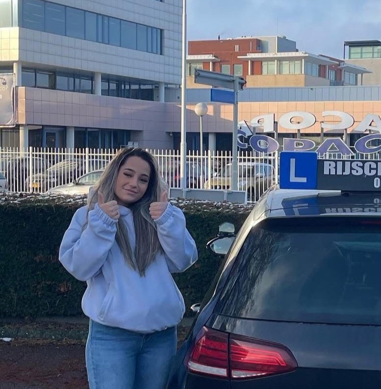 Jonge vrouw in een witte trui geeft duimen omhoog naast een auto met een 'L' leerbordje, voor een gebouw en metalen hek.