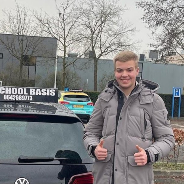 Jonge man in een grijze jas die duimen omhoog steekt naast een zwarte Volkswagen met een TAXI School Basis dakbord.
