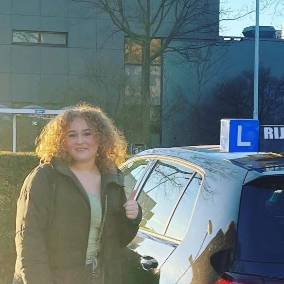 Jonge vrouw met krullend haar staat glimlachend naast een zwarte auto met een blauw 'L' rijlesbord op het dak.