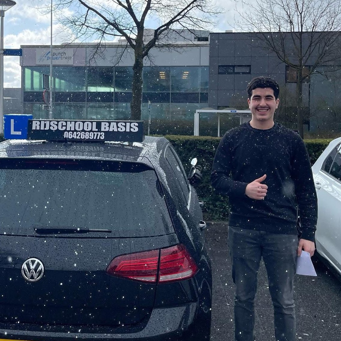 Vrouw staat lachend voor een zwarte lesauto met het bord 'Rijschool Basis' en steekt duim omhoog terwijl ze een document vasthoudt.